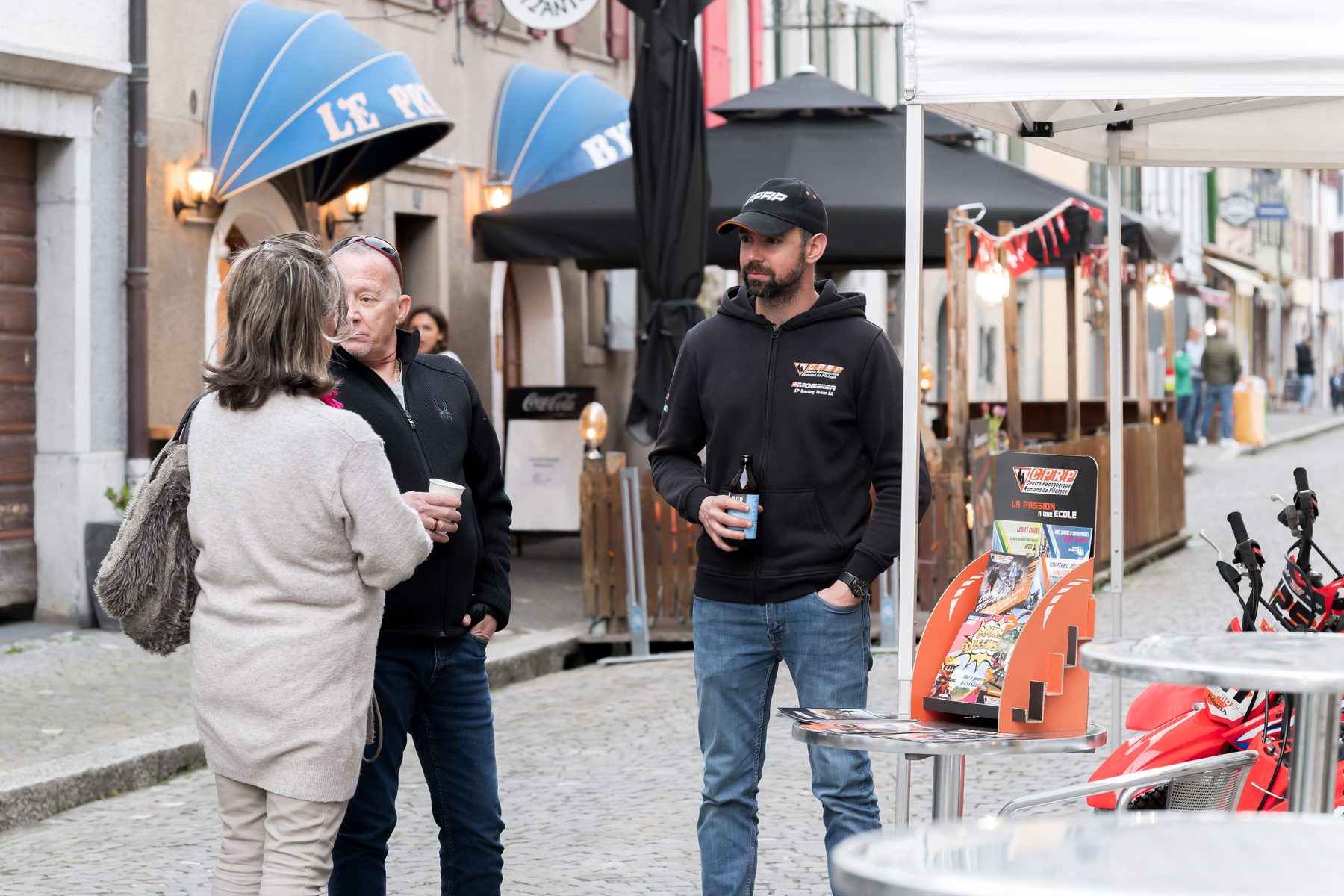 Trois adultes échangent devant un stand de motos dans la Grand’Rue
